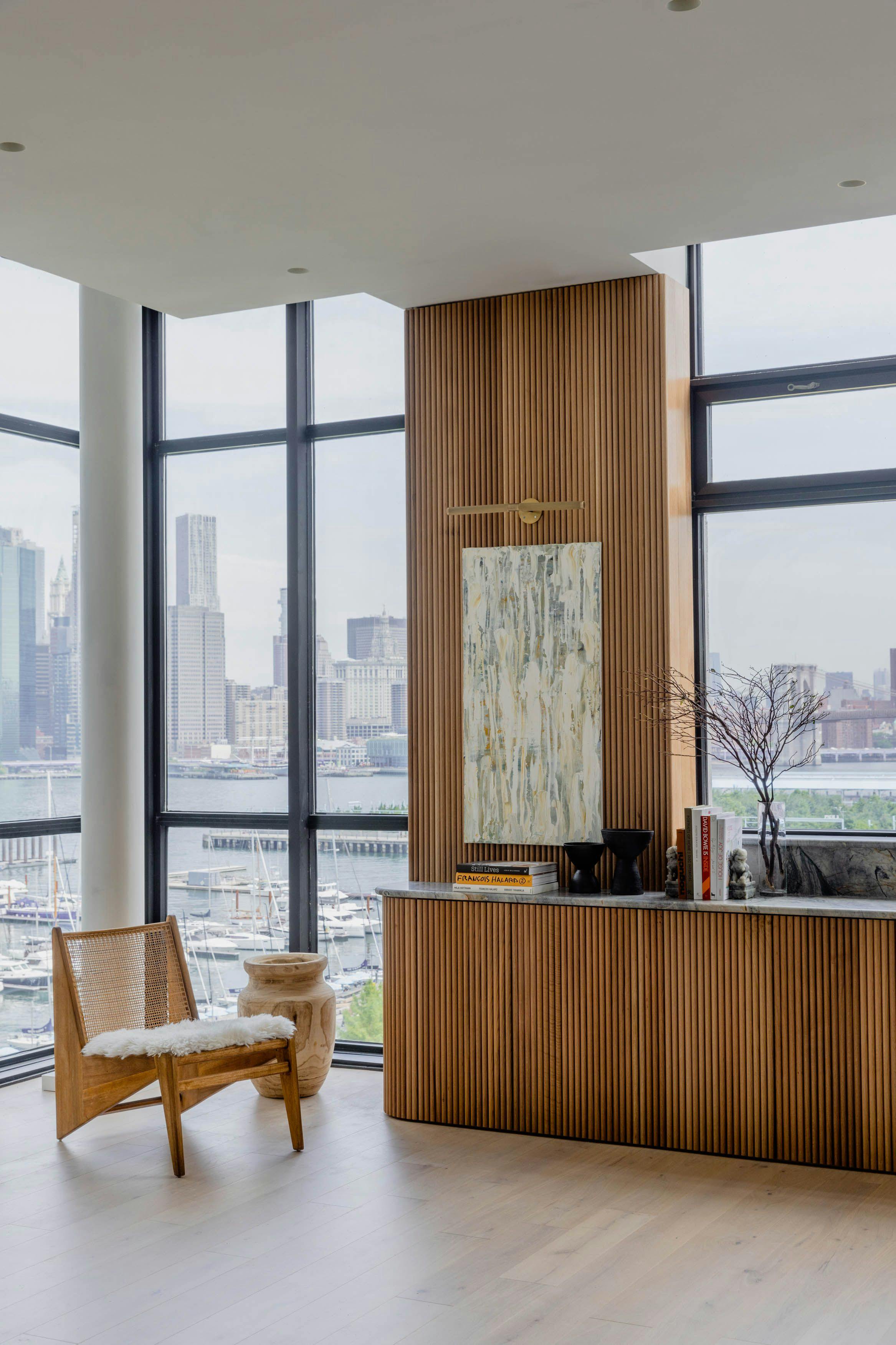 A modern living room with a city view, featuring minimalist design elements and a monolithic kitchen island overlooking the Brooklyn Bridge.